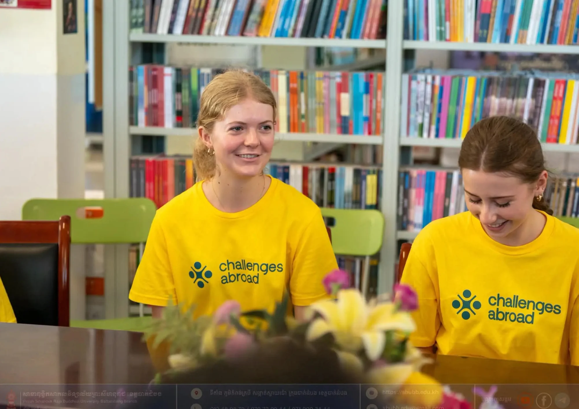 Two smiling young women in yellow 'Challenges Abroad' shirts sit in a vibrant library setting filled with colorful books.