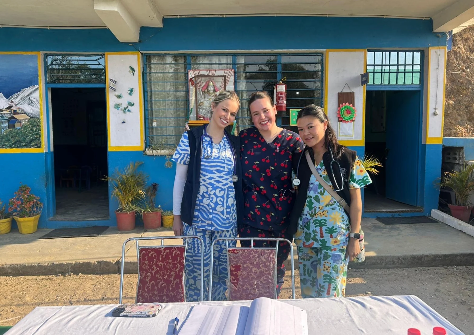 Three smiling female medical professionals in colorful scrubs pose outside a vibrant rural clinic, showcasing healthcare...