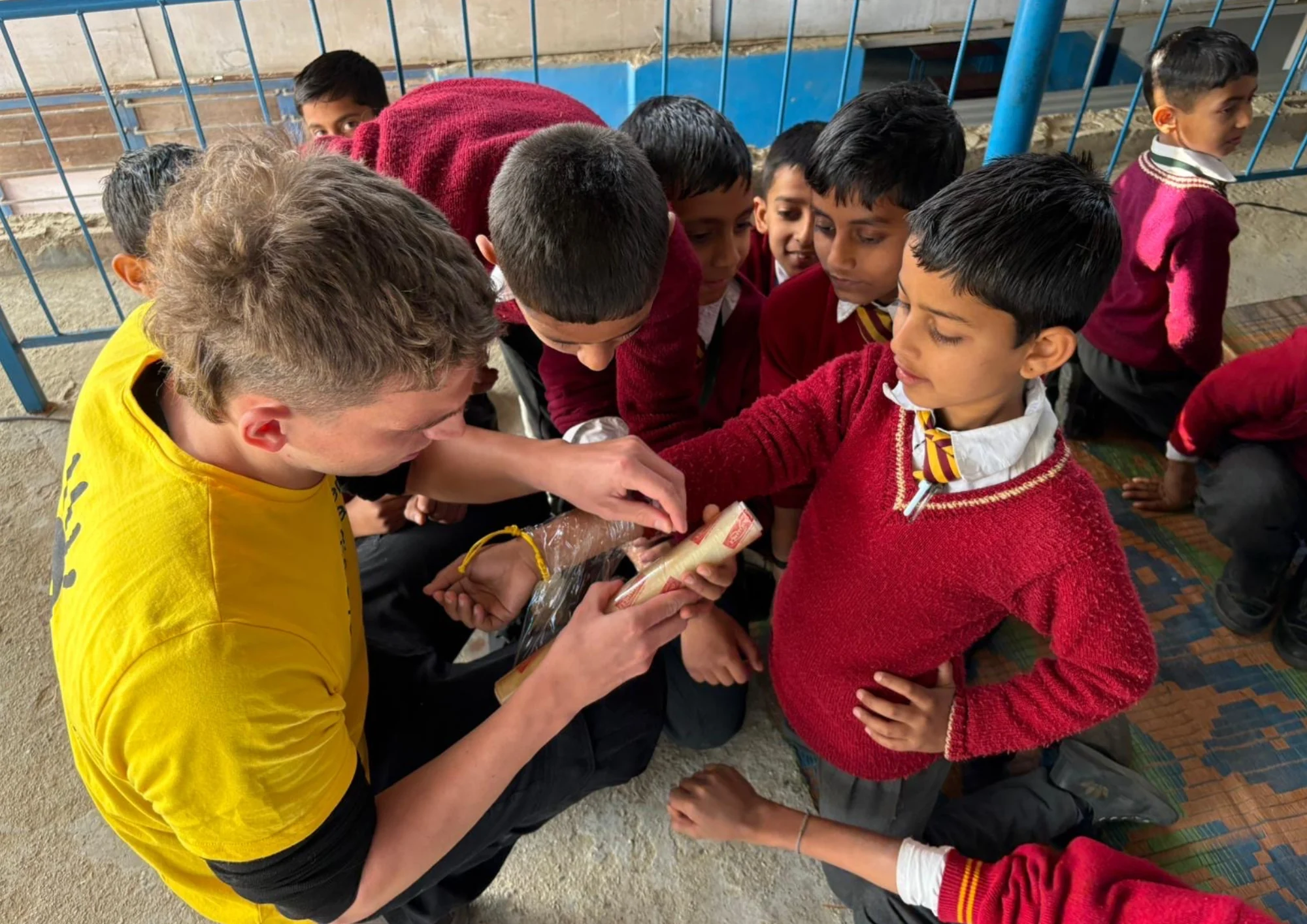 A young volunteer in a yellow shirt interacts with attentive school children wearing red uniforms, creating an engaging education setting.