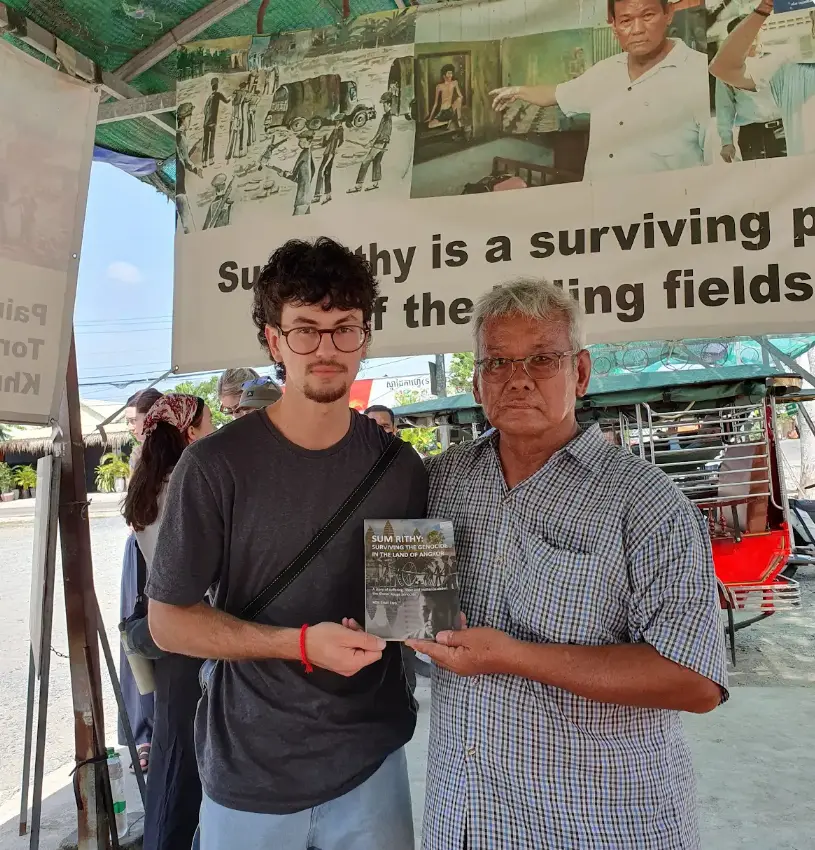Two men stand under a green tent, holding a book related to history and survival, with a banner overhead.