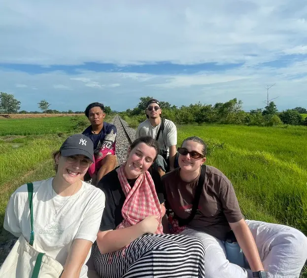 A group of friends relax on a scenic countryside rail track surrounded by lush green fields under a clear blue sky.