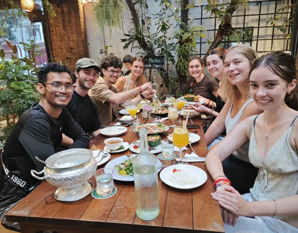 A lively group of young friends enjoy a delightful meal together in a leafy outdoor cafe setting.