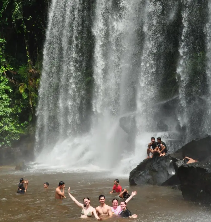 A group of people enjoy swimming beneath a stunning waterfall with lush greenery surrounding the scene.